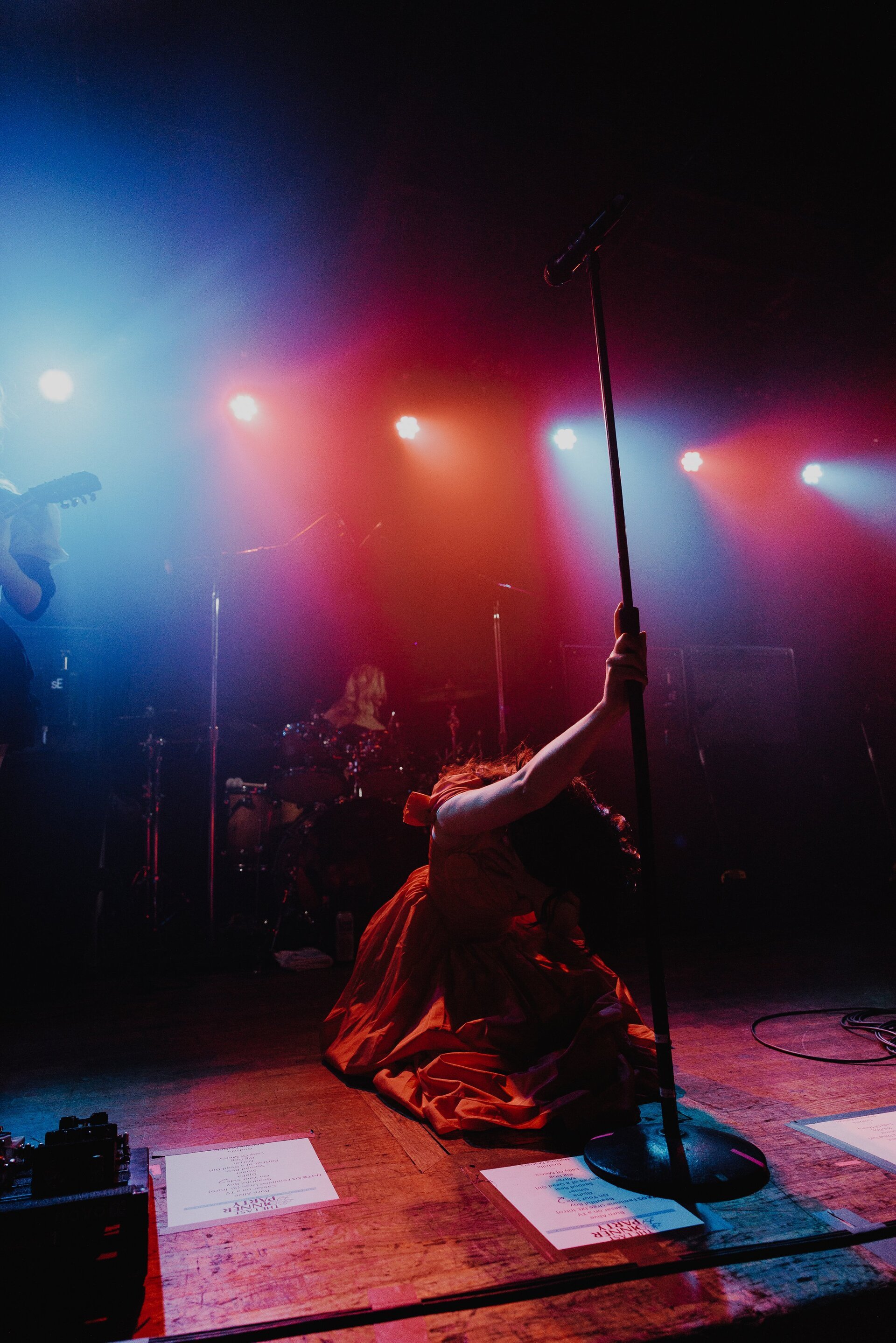 Female performer in red dress kneels, holding microphone stand on dimly lit stage with colorful spotlights during live music concert.