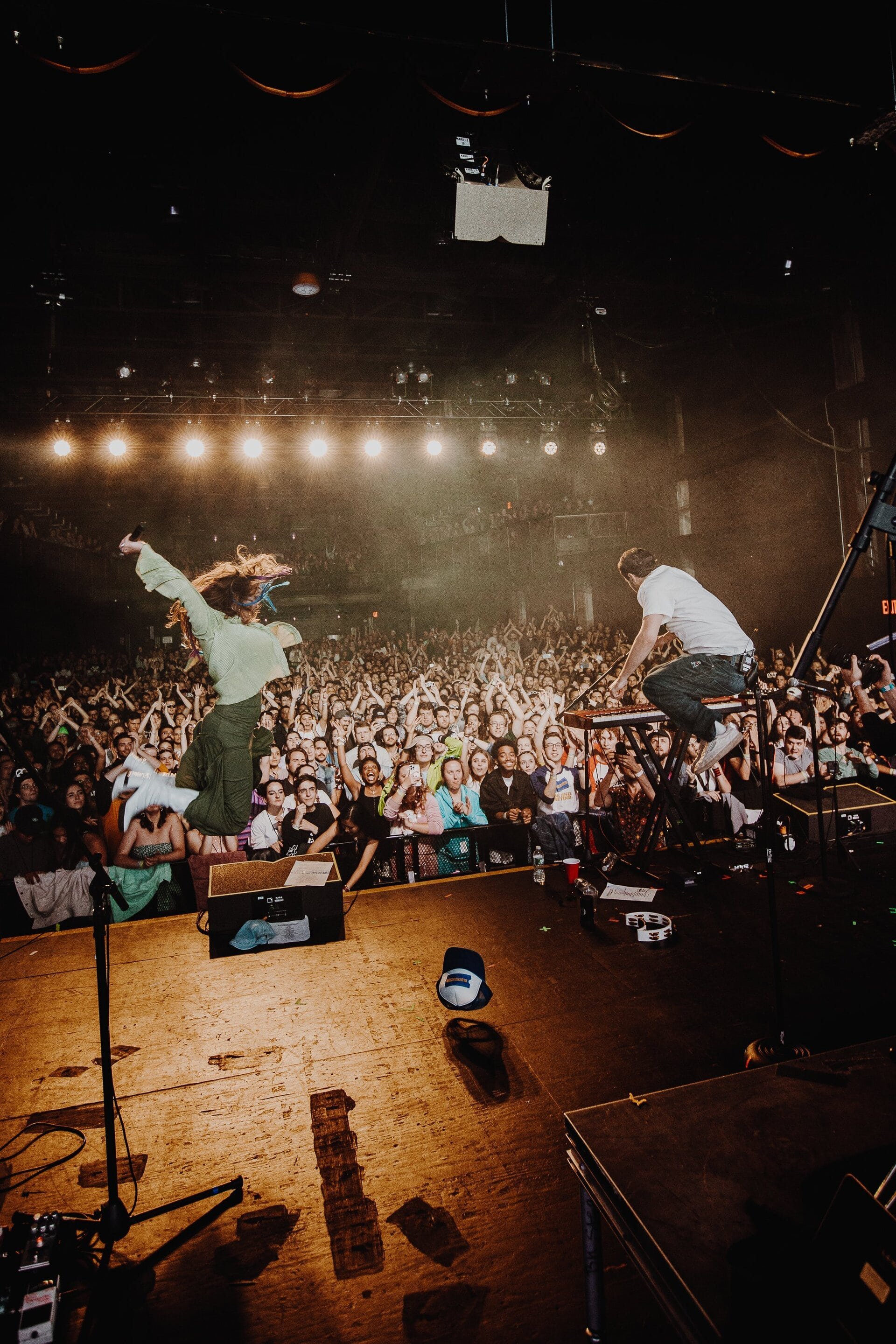Concert stage with a female musician jumping mid-air, a male musician seated, and a large crowd of people in the background.