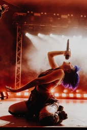 A female vocalist with blue hair kneeling on stage, leaning back while singing into a microphone under dramatic red stage lights.