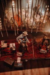 High-angle concert photography of a musician playing acoustic guitar seated on stage with a drum kit and bandmates.