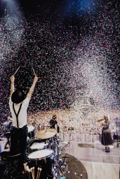 Drummer on stage with arms raised as confetti falls over a packed concert crowd, captured from behind
