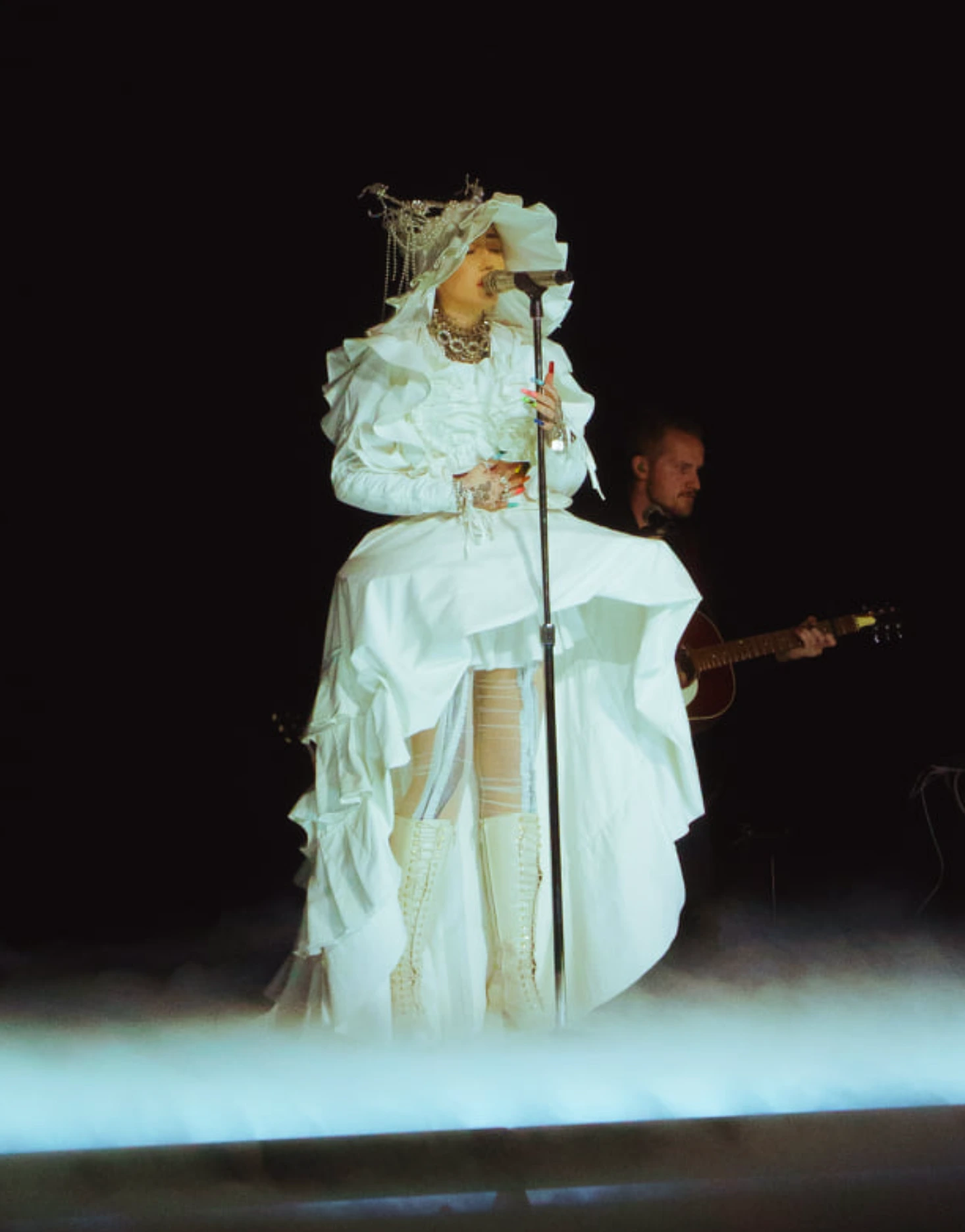 A woman in a white ruffled gown sings into a microphone on stage with a guitarist in the background, set against a dark background.