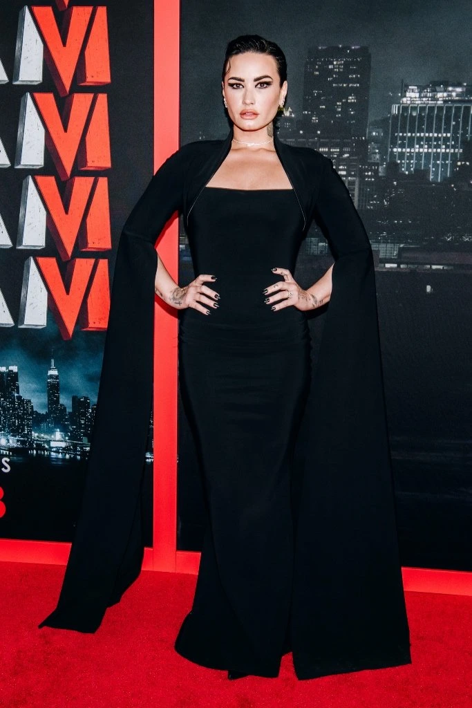 Demi Lovato poses in a black gown with flowing sleeves on a red carpet, cityscape backdrop, and vibrant event ambiance.