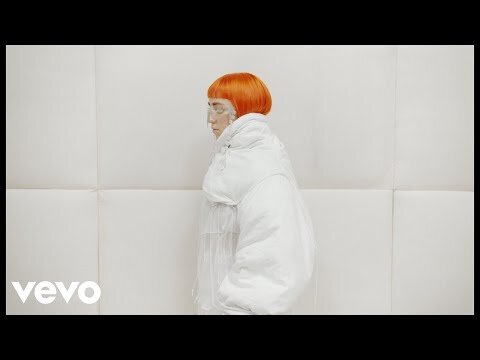 Woman with orange hair wearing a white puffy jacket in a minimalist white photo studio with a large sheet of white paper backdrop.