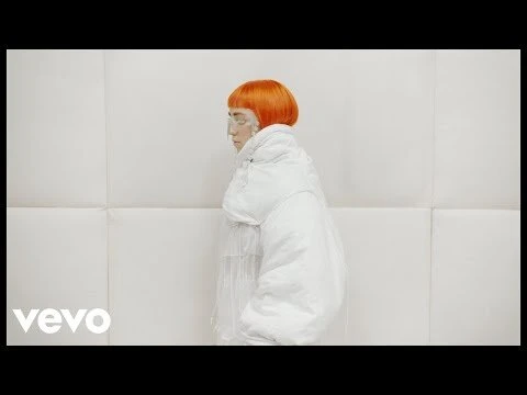 Woman with orange hair wearing a white puffy jacket in a minimalist white photo studio with a large sheet of white paper backdrop.