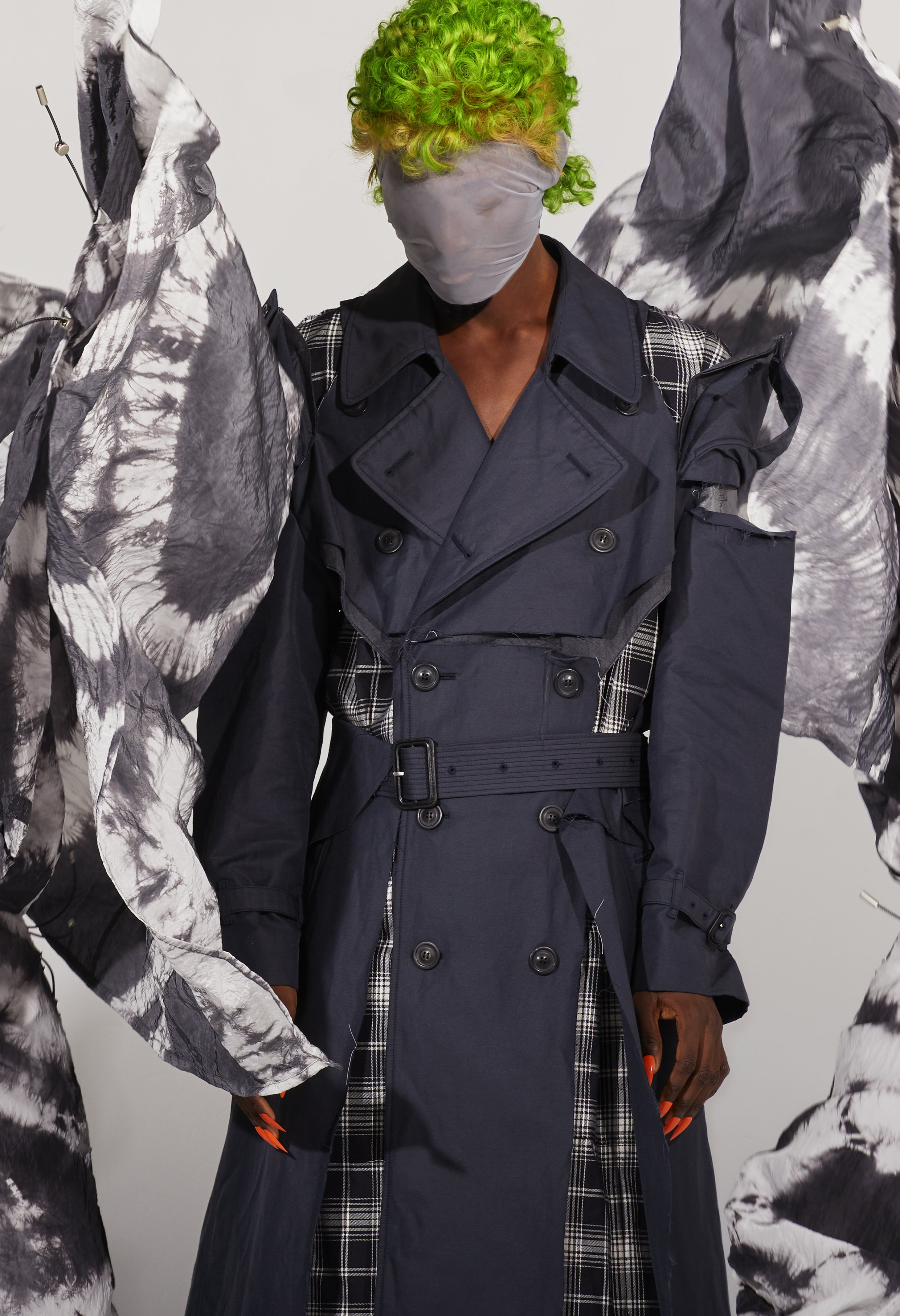 Fashion model with green hair wearing deconstructed navy trench coat and head covering, standing in studio with decorative fabric.