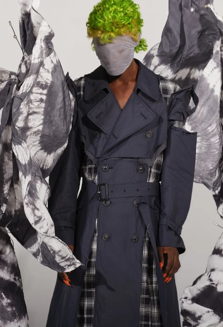 Fashion model with green hair wearing deconstructed navy trench coat and head covering, standing in studio with decorative fabric.