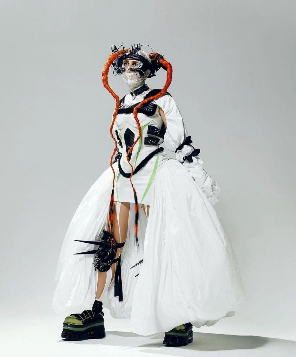 Fashion model in a surreal, avant-garde outfit, featuring orange hair extensions, platform shoes, and a theatrical white dress, studio shot.