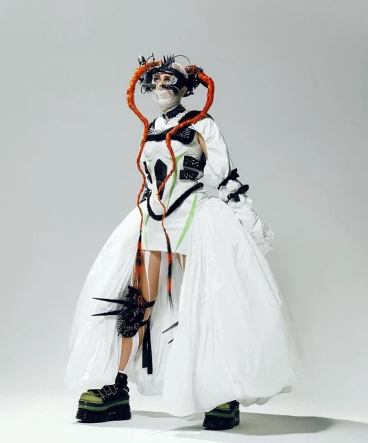 Fashion model in a surreal, avant-garde outfit, featuring orange hair extensions, platform shoes, and a theatrical white dress, studio shot.