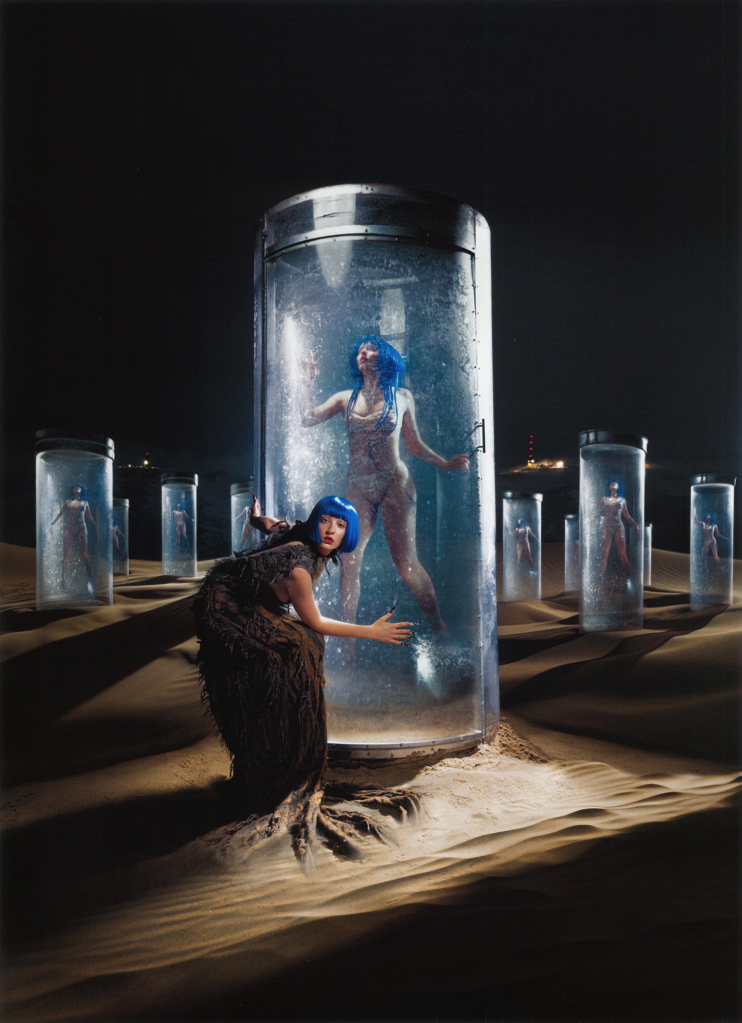 Two women with blue wigs, one standing in a cylinder of water and the other leaning forward in the sand, stand in a sandy landscape.