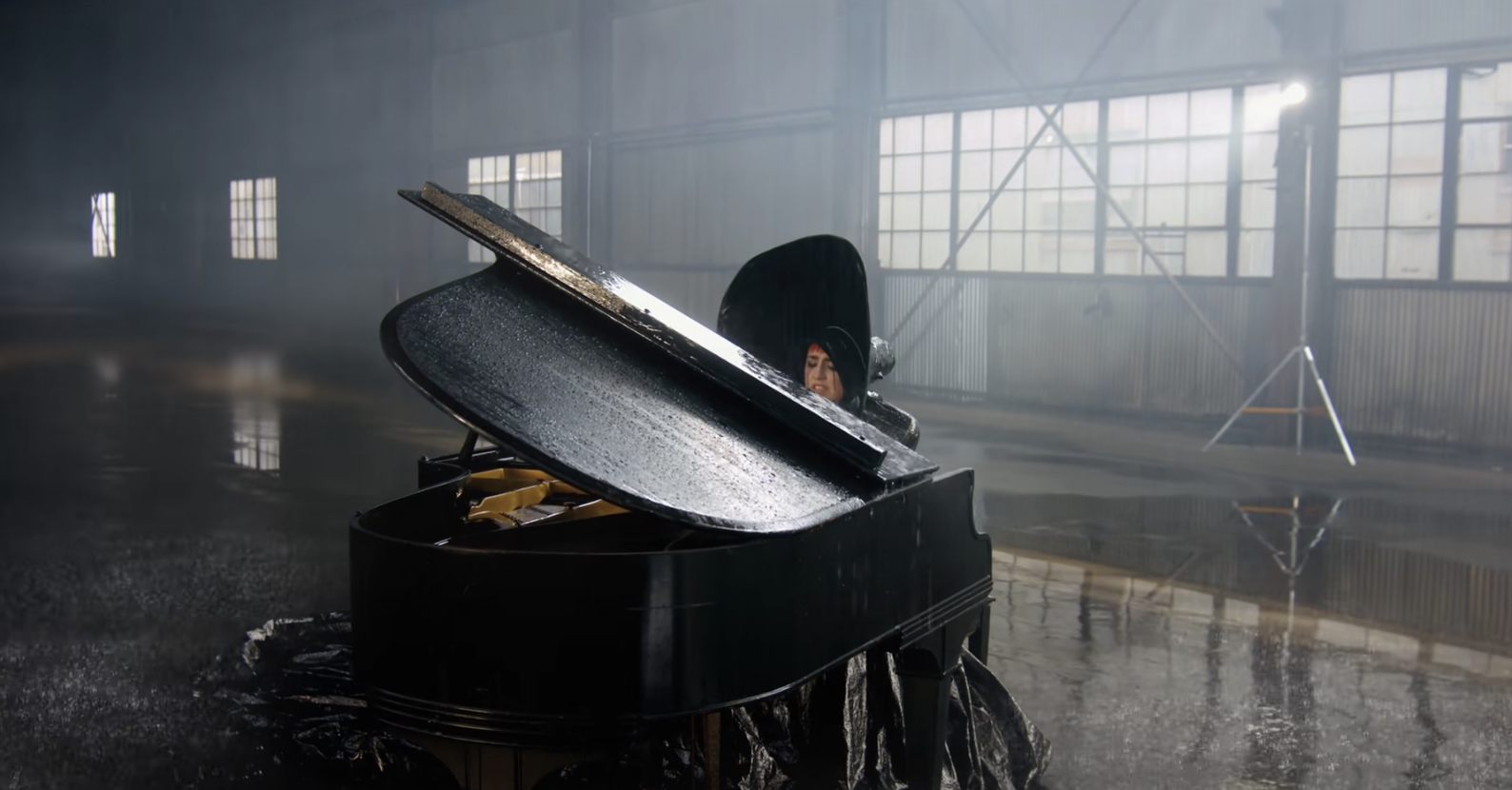 A woman sits behind an open grand piano in a large, empty hall with a wet floor, illuminated by stage lighting.