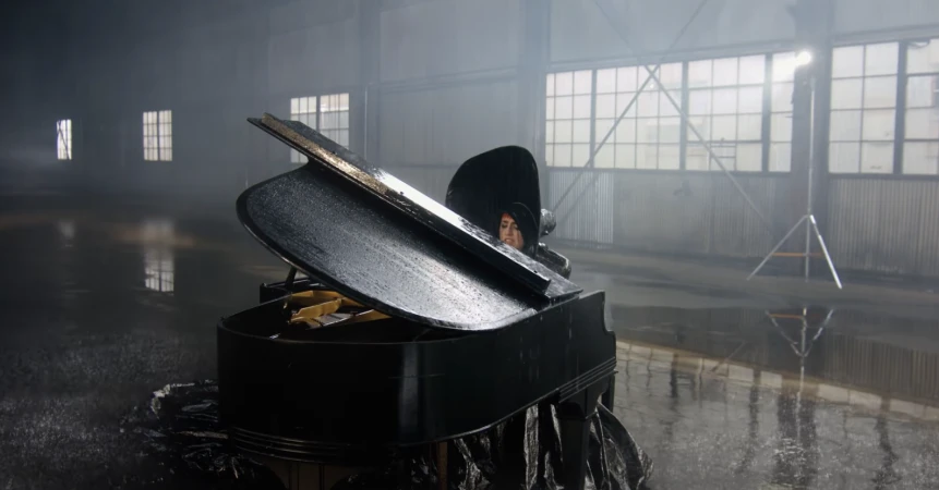 A woman sits behind an open grand piano in a large, empty hall with a wet floor, illuminated by stage lighting.