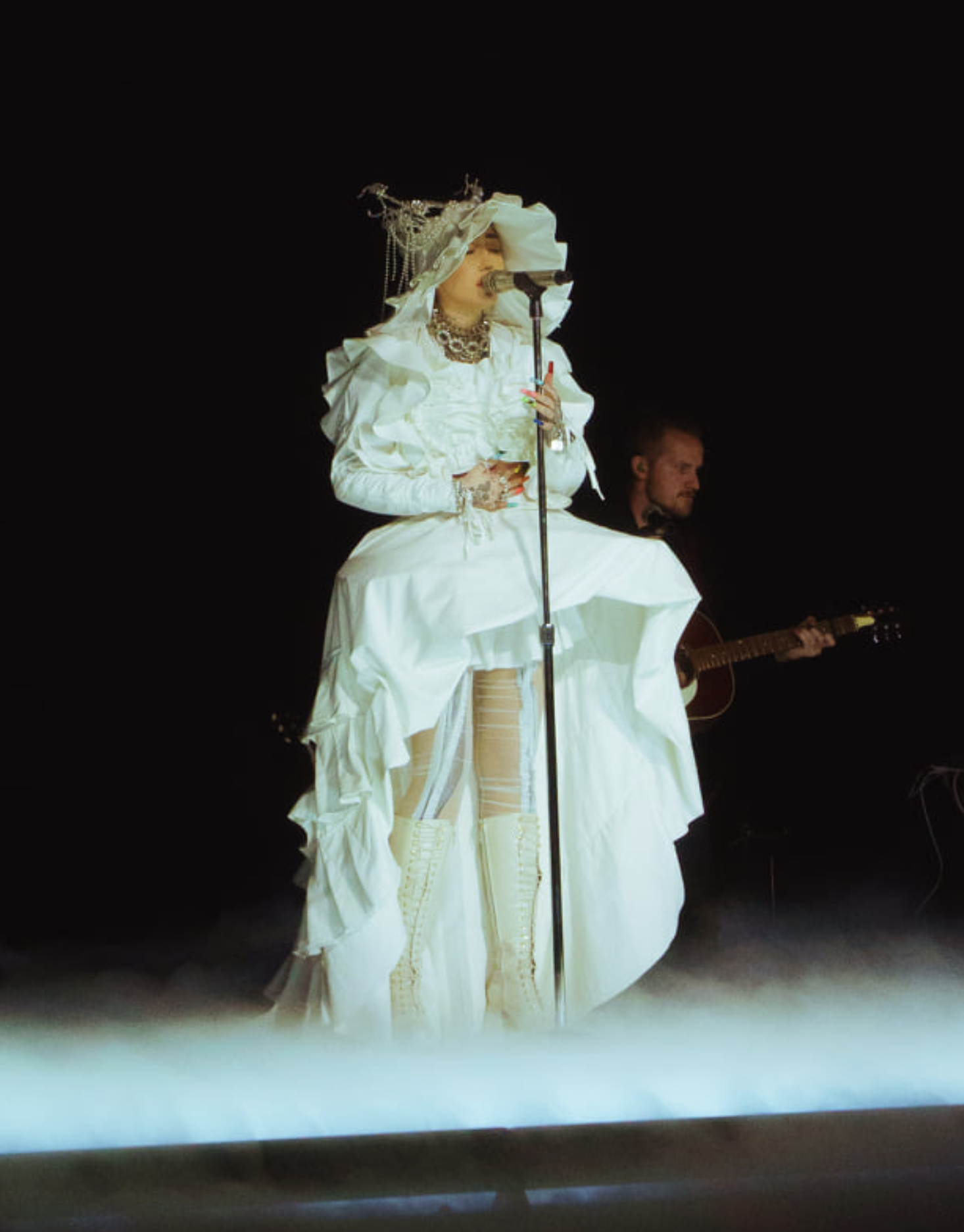 Female singer in a white costume performs on stage with a guitarist in the background against a black backdrop and stage lights.