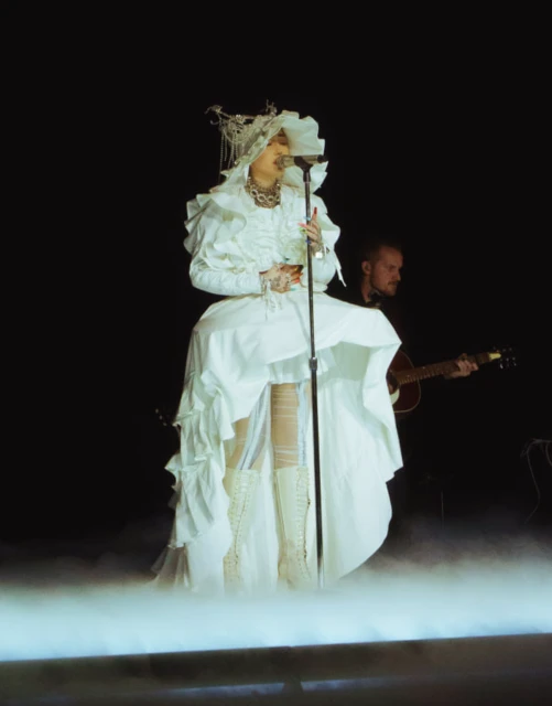 Female singer in a white costume performs on stage with a guitarist in the background against a black backdrop and stage lights.