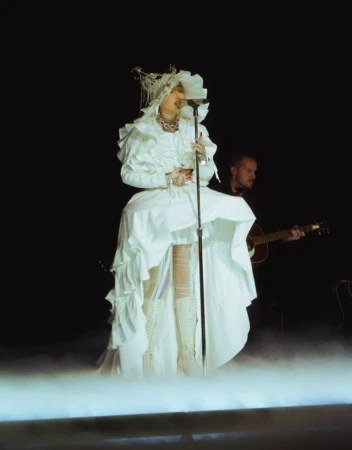 Female singer in a white costume performs on stage with a guitarist in the background against a black backdrop and stage lights.