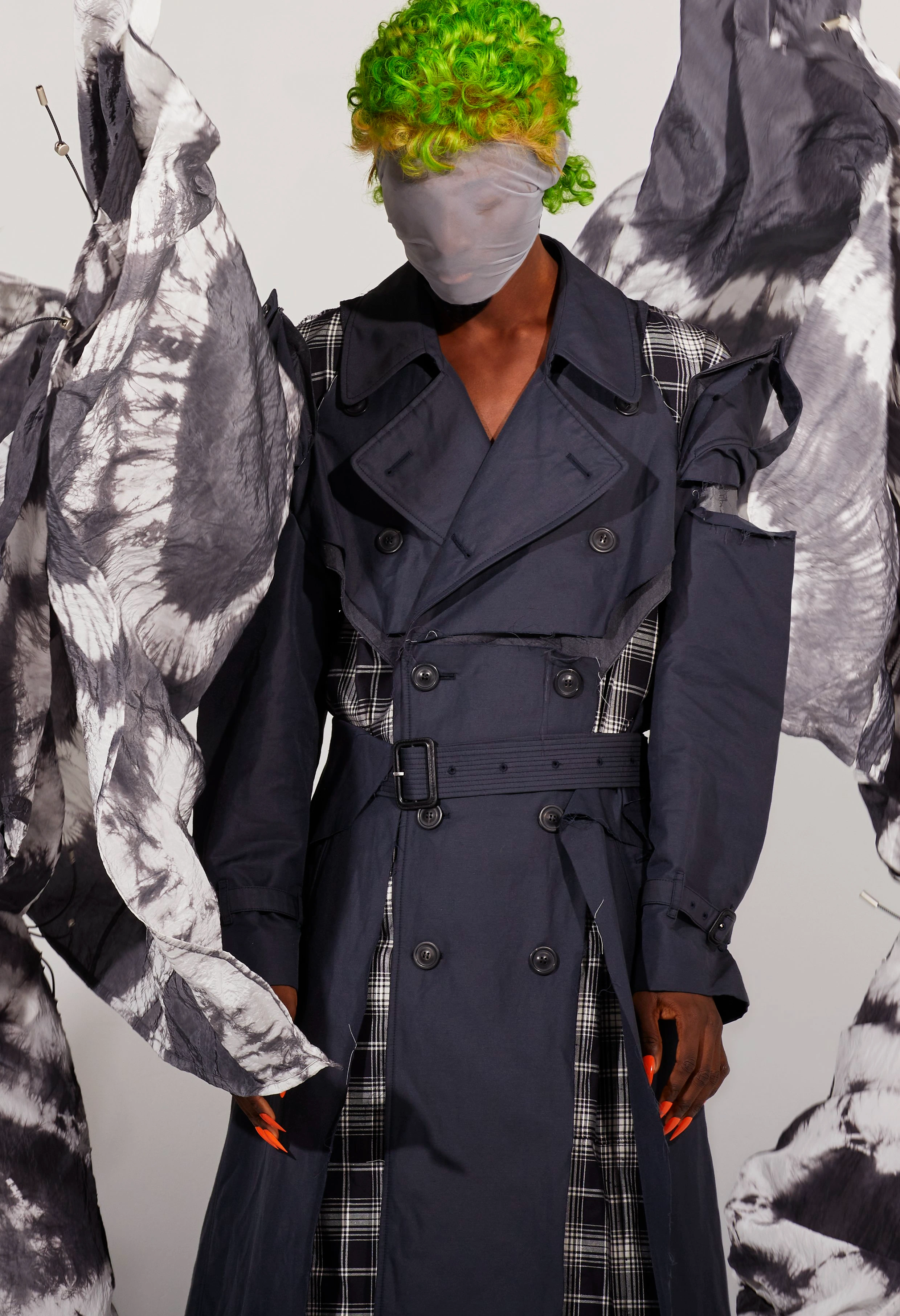 Model in a navy trench coat with plaid details and green hair, posing in a studio. Fashion editorial with unique details and muted colors.