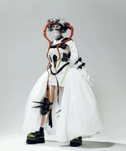 Unique fashion portrait of a person wearing a distinctive white dress with platform shoes against a gradient grey studio background.