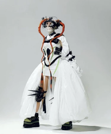 Unique fashion portrait of a person wearing a distinctive white dress with platform shoes against a gradient grey studio background.