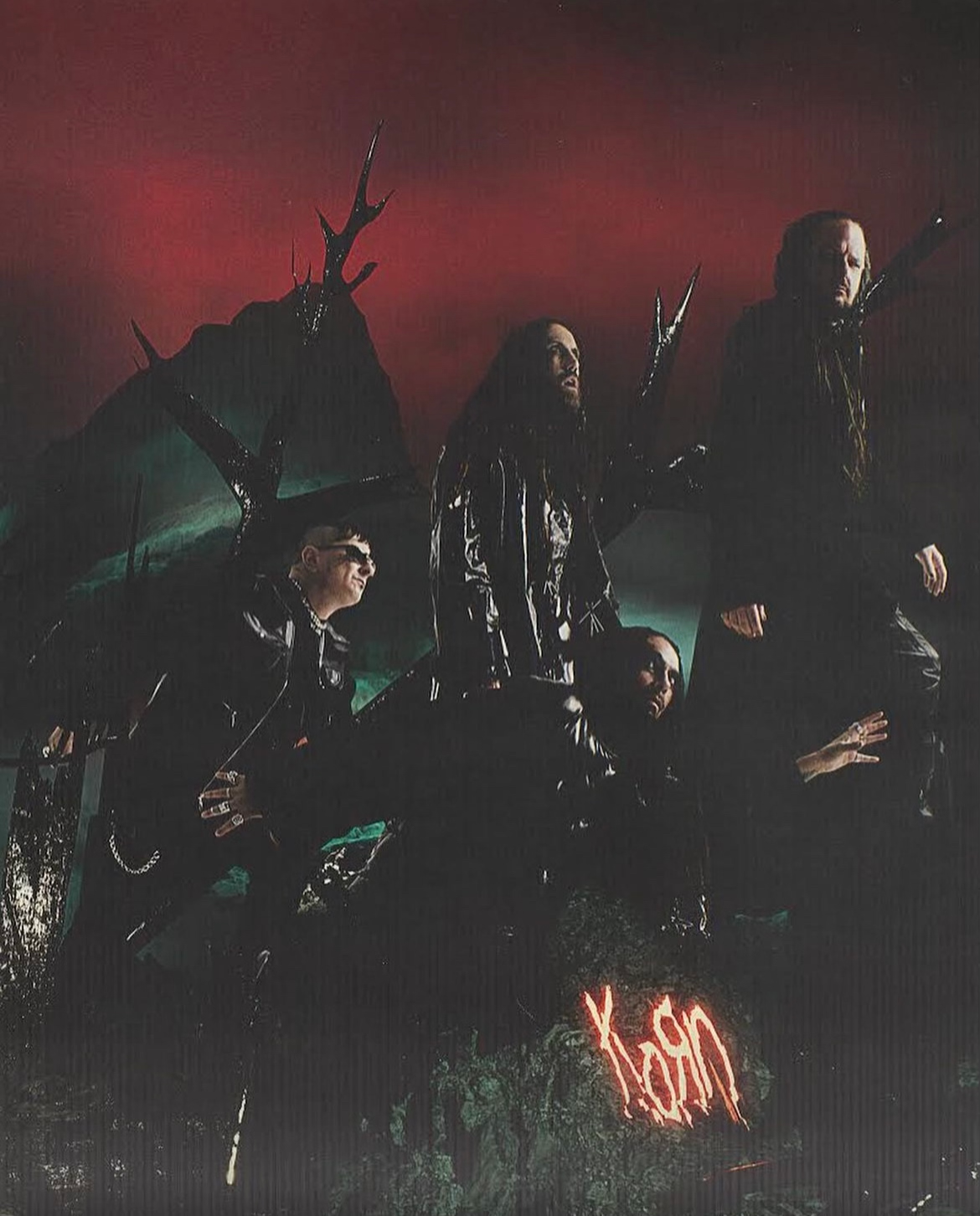Band members of Korn in dark clothing stand against a dark forest backdrop with a red sky, illuminated by neon writing.