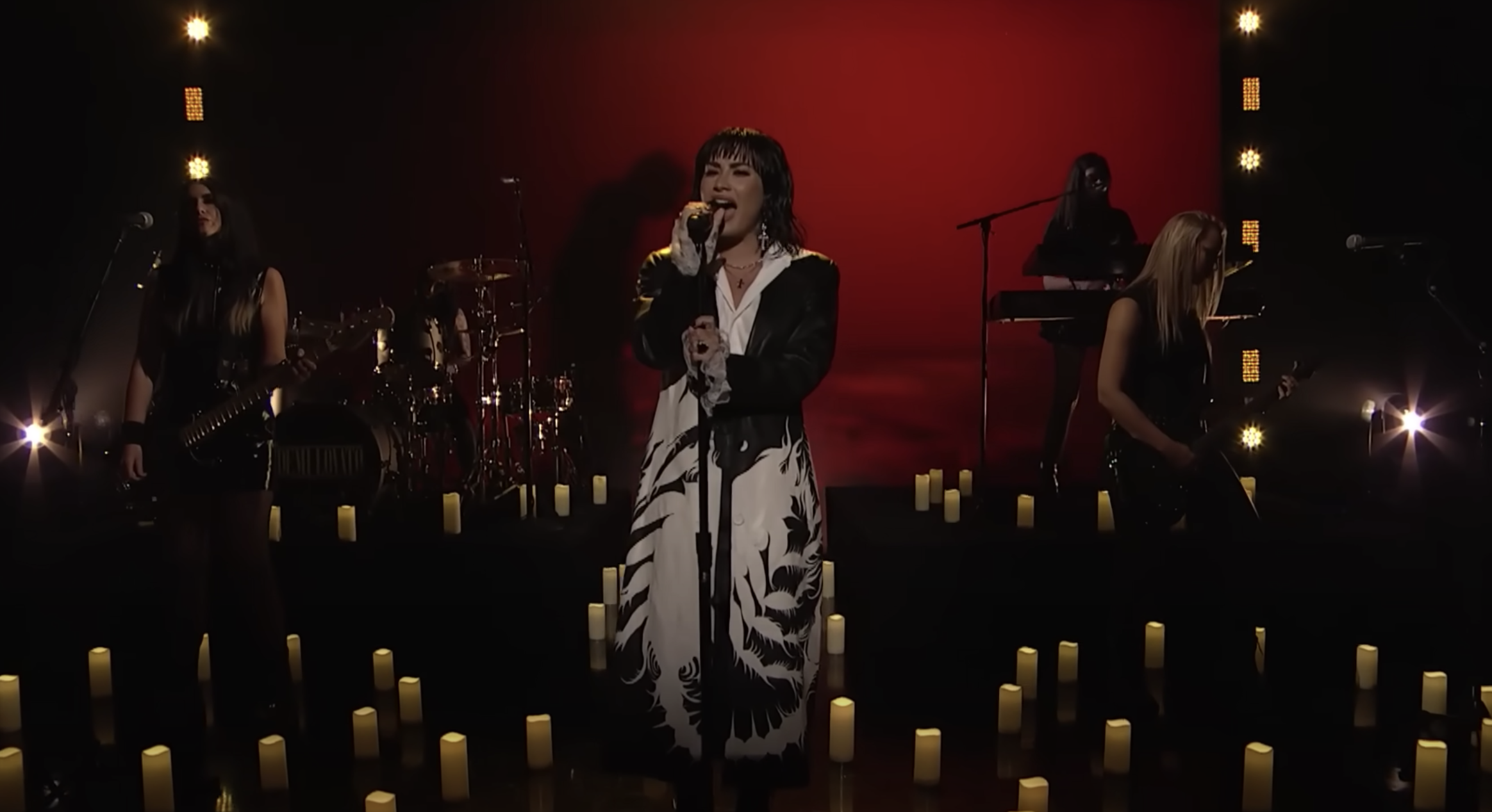 Female singer performing on stage with her band in dim lighting, surrounded by candles and a red backdrop during a live music event.
