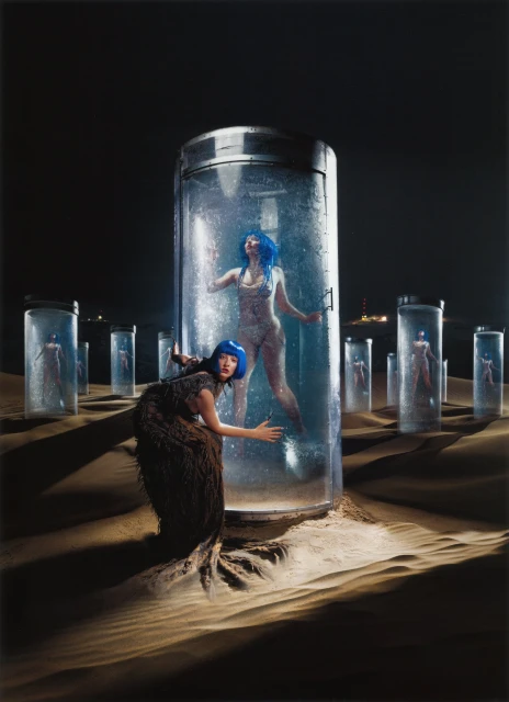 Two women with blue hair near glass containers in a desert setting at night; one woman is inside a water-filled container.