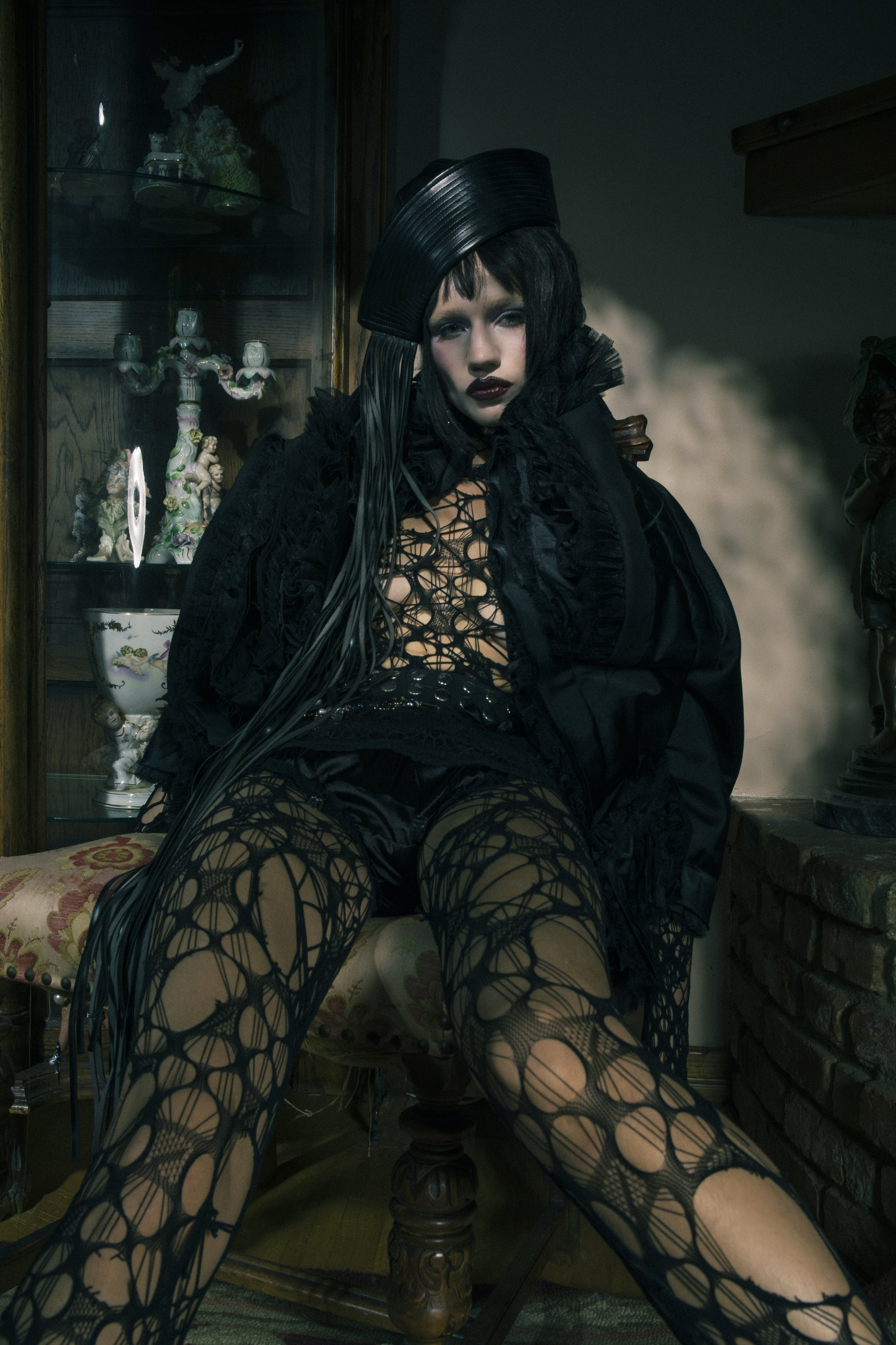 A dark portrait of a person with pale skin and dark makeup sitting in a chair wearing a hat and black, patterned outfit, indoors.