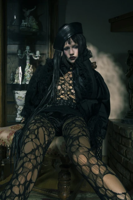 A dark portrait of a person with pale skin and dark makeup sitting in a chair wearing a hat and black, patterned outfit, indoors.