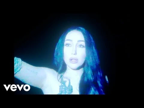 Close-up portrait of a singer bathed in blue lighting with a slight smile, set against a dark background during a music video recording.