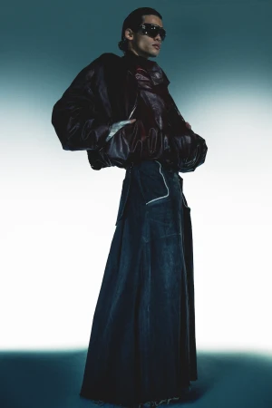 Model wearing dark sunglasses, an oversized leather jacket, and a long dark skirt against a bright gradient background by Maleeka Moss.