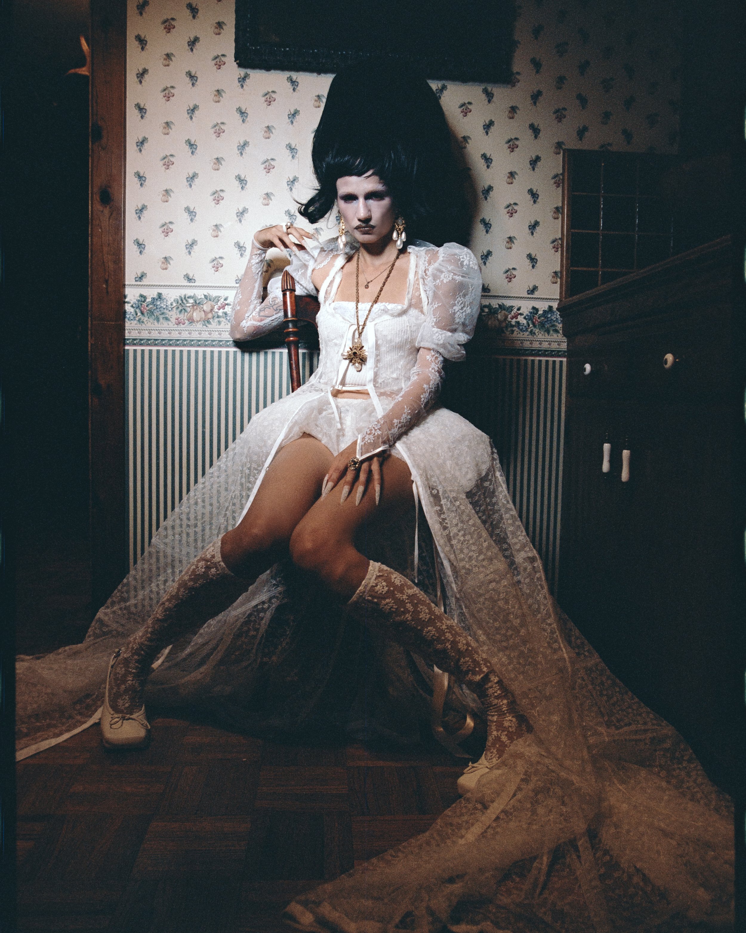 Model in a lace dress and large wig sitting on a chair. Indoor studio photo with a patterned wall and hardwood floors.