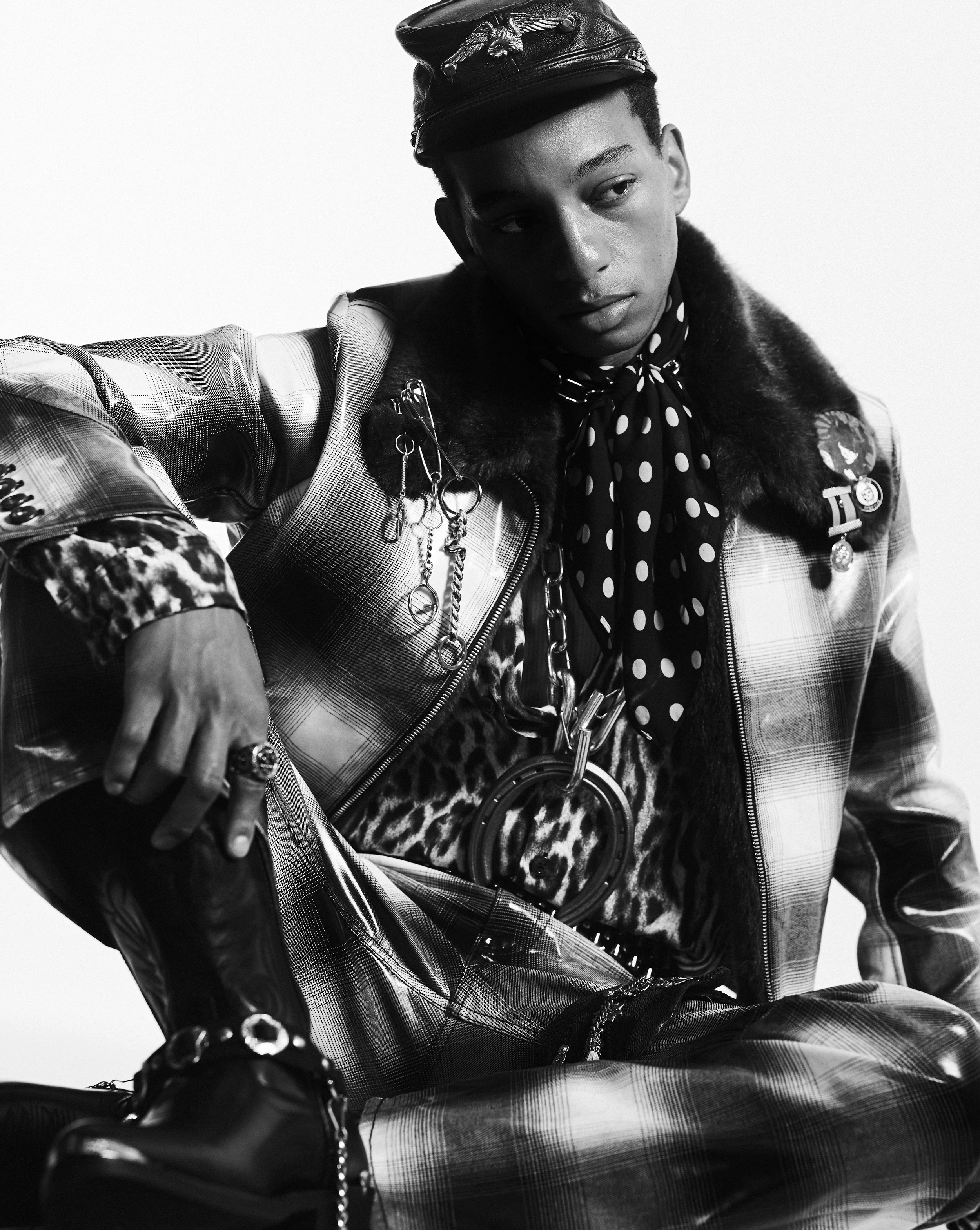 Portrait of a male fashion model with a cap and jewelry. He is wearing a plaid jacket, a leopard-print shirt, and black leather shoes.