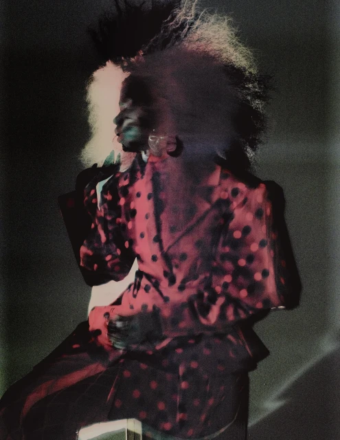 Woman with afro hairstyle, wearing a red and black polka dot blazer, posing in a dark studio setting with lighting effects.