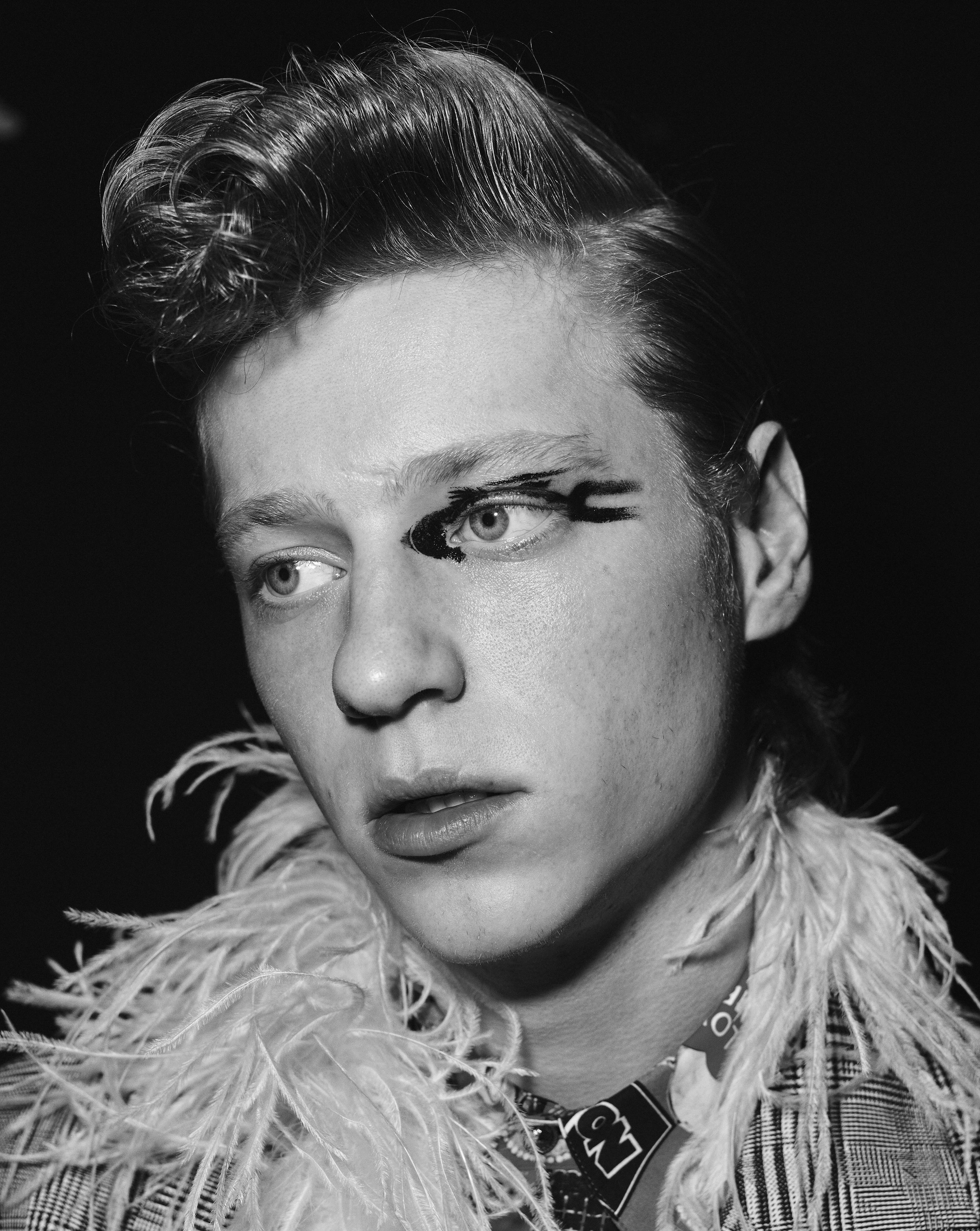 Black and white portrait of a young adult male with stylized black eye makeup, wearing a feathered collar and patterned shirt.