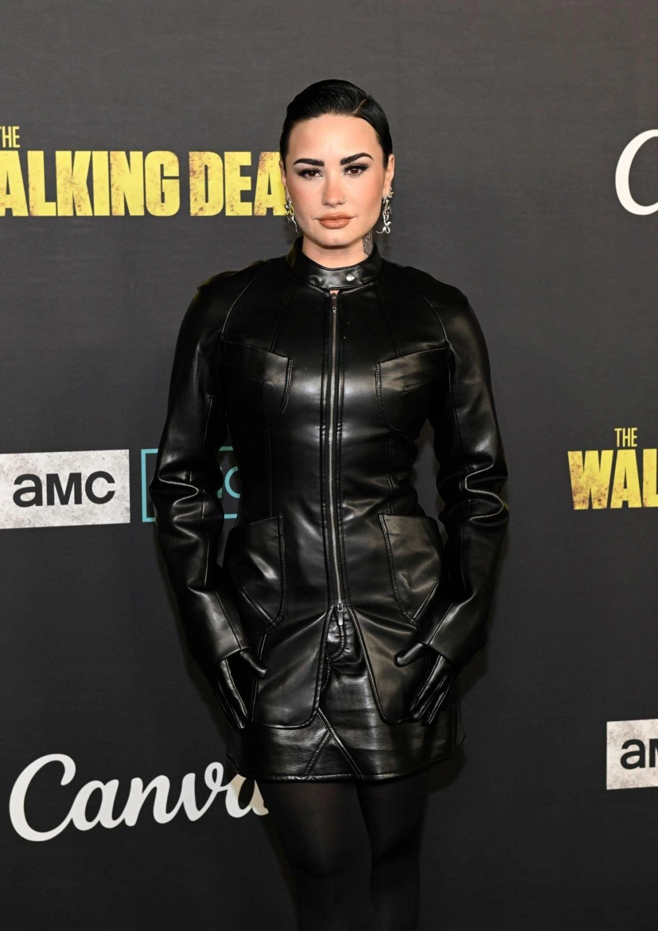 Demi Lovato at an event for The Walking Dead. She is wearing a black leather dress and gloves and is posing for the camera.