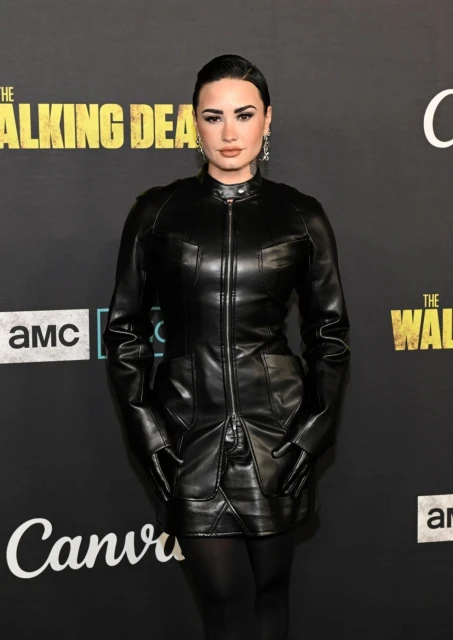 Demi Lovato at an event for The Walking Dead. She is wearing a black leather dress and gloves and is posing for the camera.