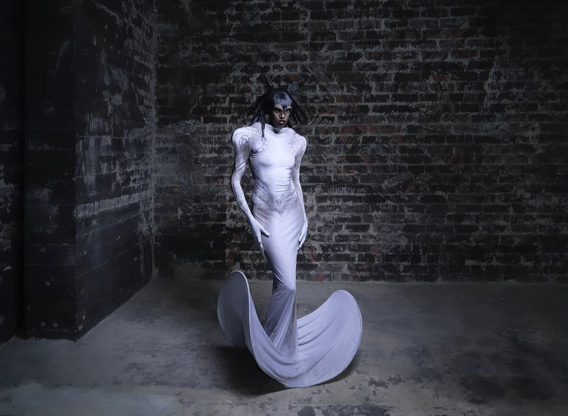 Woman in a white sculptural gown poses against a brick wall in an indoor setting, creating a dramatic fashion image with unique style.