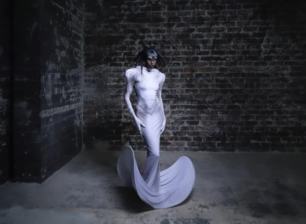 Woman in a white sculptural gown poses against a brick wall in an indoor setting, creating a dramatic fashion image with unique style.