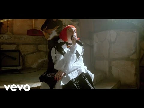 Singer with red hair performs on a stone stage with a musician, bathed in warm lighting, creating a captivating musical scene.