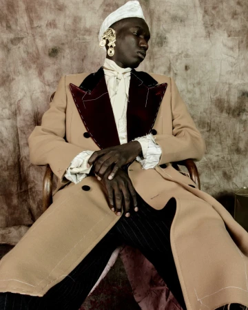 Portrait of a Black man in a beige coat and a white head covering sitting in a wooden chair against a neutral mottled backdrop.