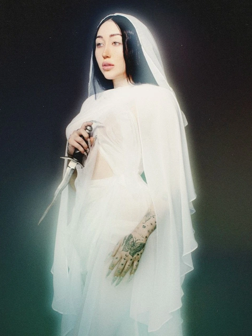 Portrait of a young woman in an ethereal white gown and veil holding a dagger in a dimly lit, moody setting, showcasing dark hair.