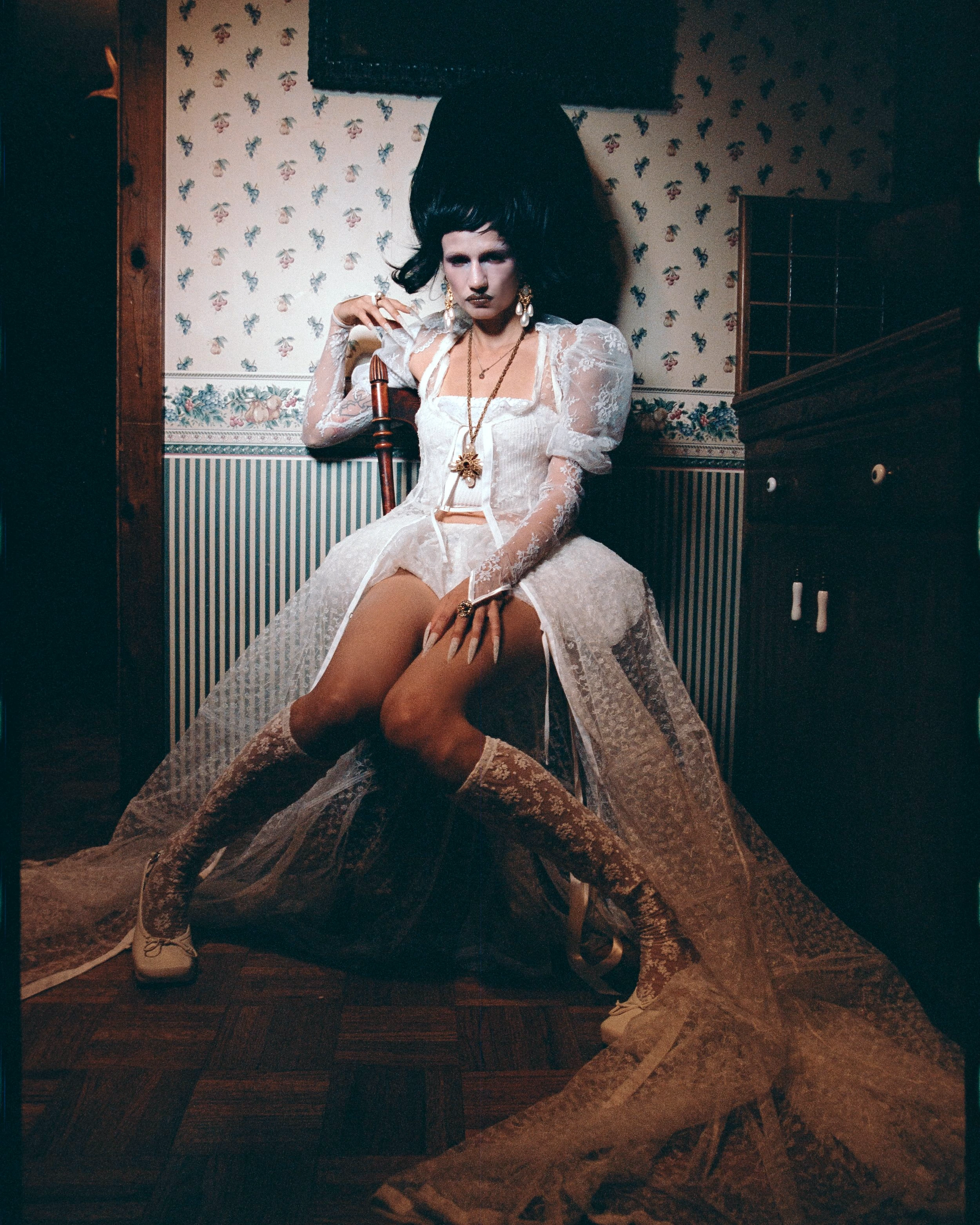 A person with a large afro wig poses in a white lace gown, boots, and lingerie. The image is indoors with patterned wallpaper and wood floors.