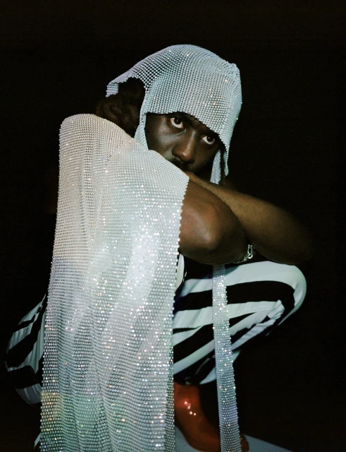 Stylish portrait of a Black adult male crouching in a shimmering head covering, wearing black and white striped pants against a dark backdrop.