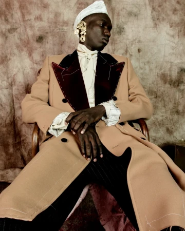 A Black man seated in a wooden chair wearing a tan coat with dark lapels, a white ascot, and large gold earrings.