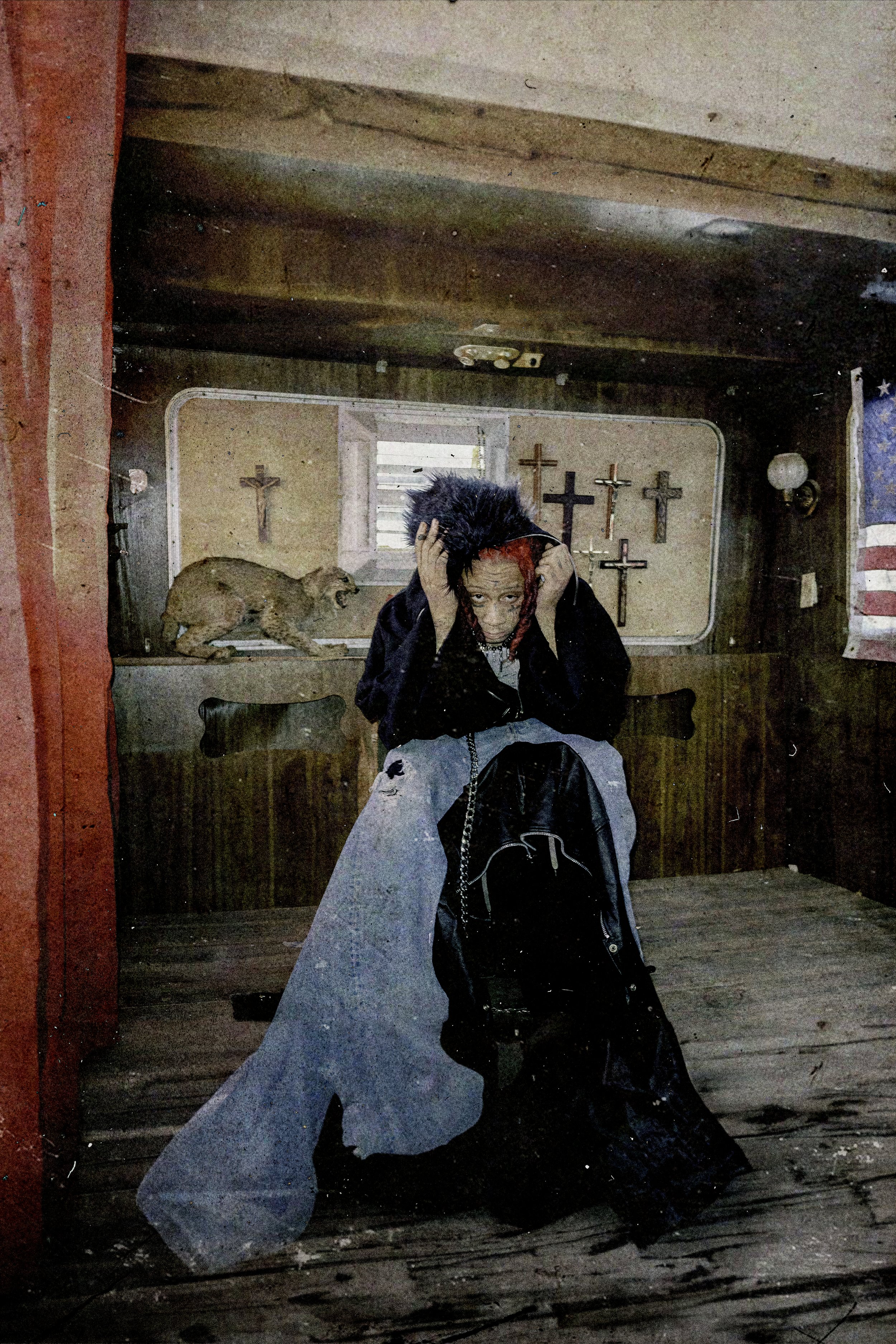 An adult female sits in an aged interior space with wood paneling, next to crosses and a bobcat taxidermy. She wears ripped jeans.