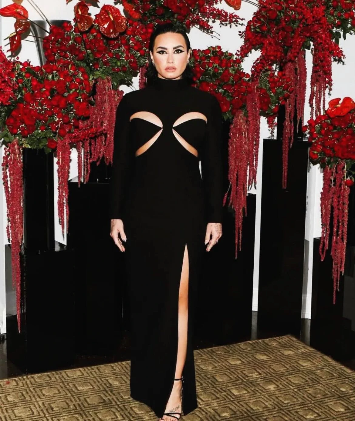 Demi Lovato in a black, long-sleeved gown with cut-out details, standing against a backdrop of cascading red floral arrangements.