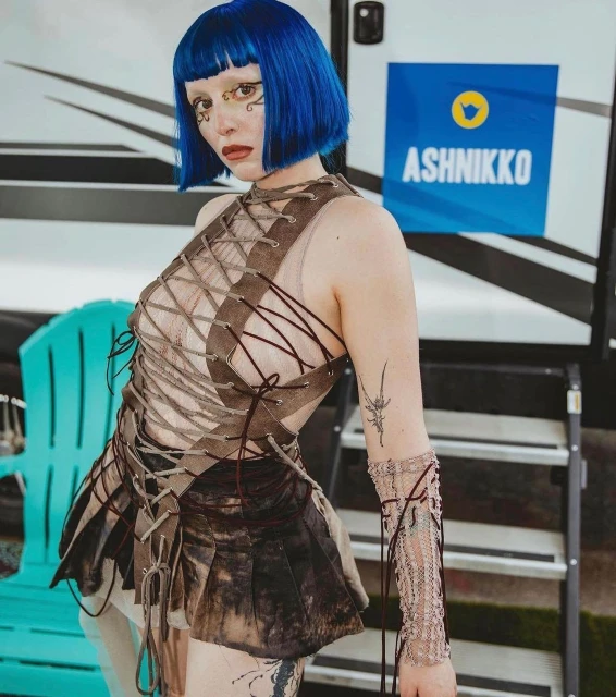 Woman with blue hair poses in a patterned brown dress with a backdrop of a trailer and turquoise chair for a fashion editorial.
