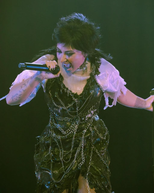 Female vocalist performing on stage wearing a black dress with chain details, singing into a microphone with dark, dramatic makeup.