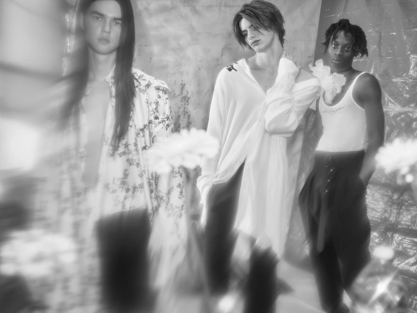 Three fashion models pose indoors under bright light and soft focus. The models wear white shirts and dark pants against a textured backdrop.