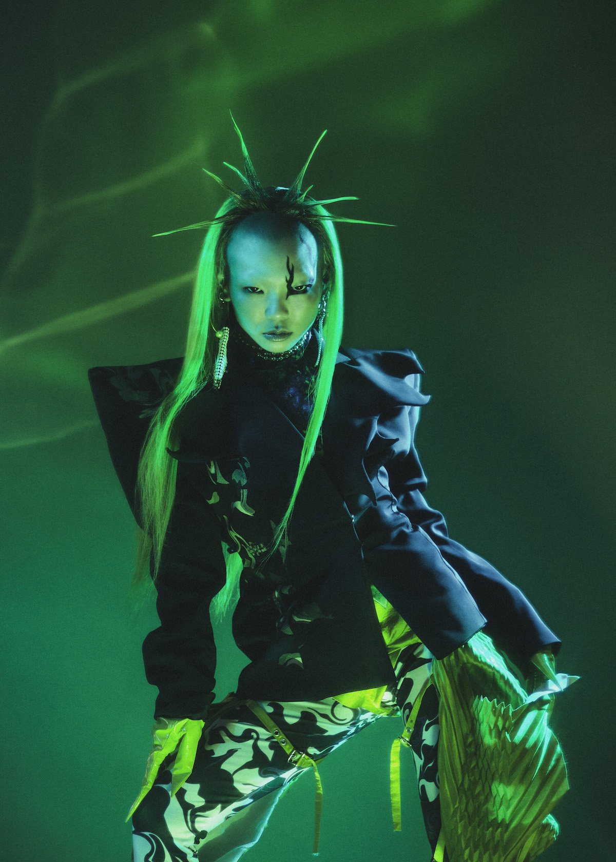 Model poses in dark jacket with neon green hair against a neon green background. Dramatic, high-fashion editorial shot.