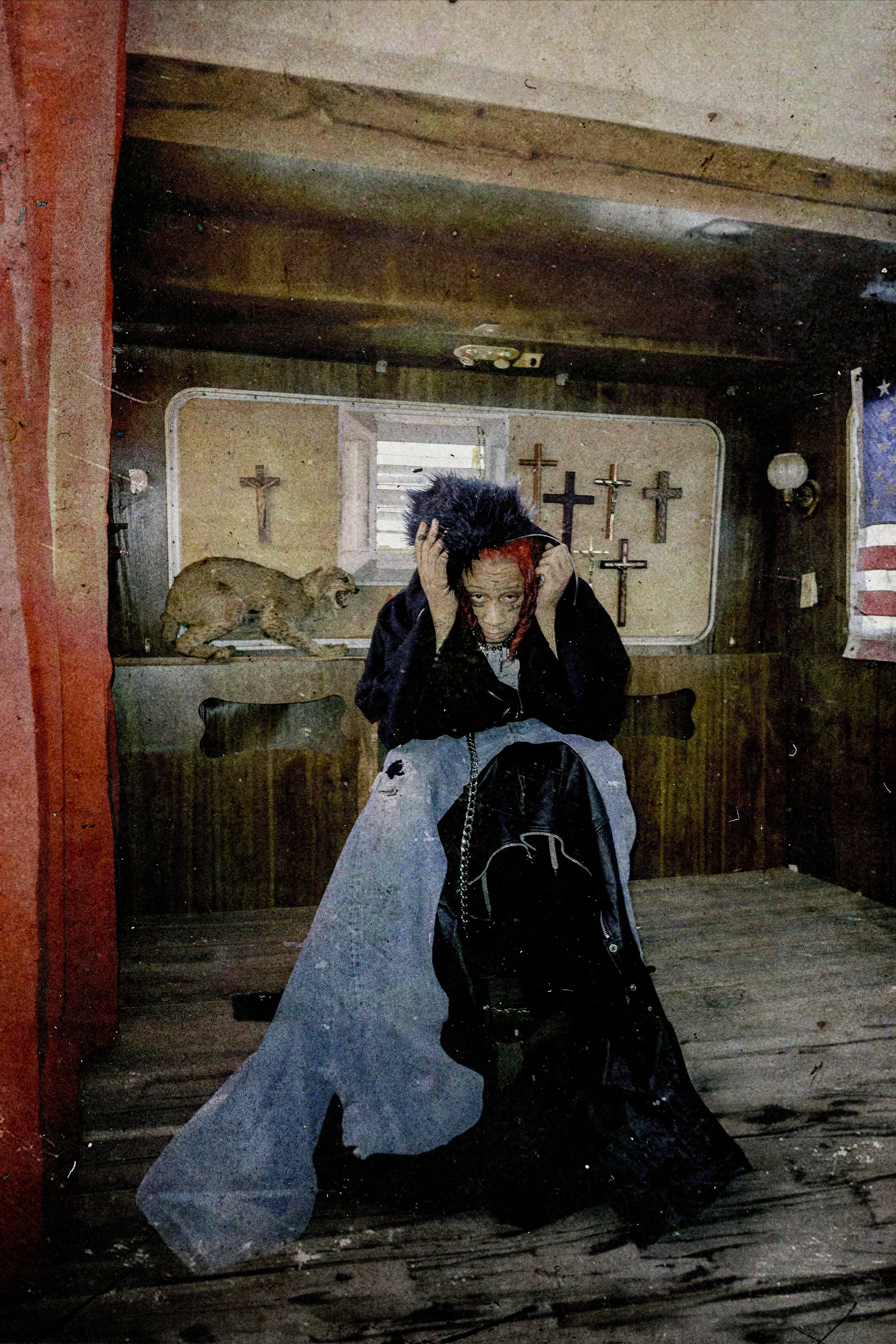 Woman with dark hair sits indoors in front of religious display, with an American flag visible, wearing black coat and blue pants.